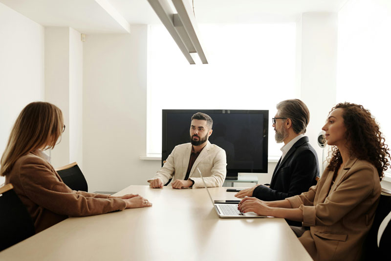 Four people are seated around a conference table in a well-lit office. One person is speaking, while the others listen attentively. A large screen is visible in the background.