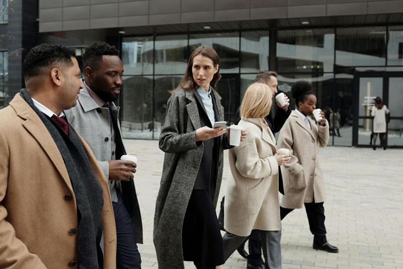 A group of six people walking outdoors in a city, each holding a coffee cup. They are dressed in formal coats, appearing engaged in conversation, with modern buildings in the background.